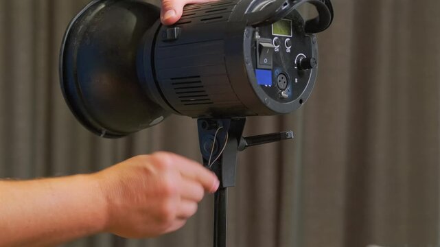 Hands mounting studio flash on light stand close-up, securing strobe unit into holder mount, preparing professional lighting equipment for photo or video production, demonstrating setup process 