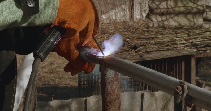 Close-up view of a welder's hands in protective gloves using an electrode to weld a metal pipe for a fence, creating bright sparks and smoke in an outdoor setting during the day.