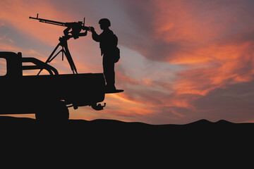 A silhouette of a soldier with a machine gun stands on a vehicle against a sunset backdrop. A concept for defending airspace against drone attacks. Themes: military, security, patrols. Defense © Andrew L