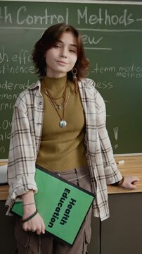 Medium portrait of 16-year-old teenage Caucasian schoolgirl standing by blackboard with chalk notes on birth control methods, while attending sexual education lesson, looking at camera with shy smile