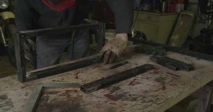 Skilled male welder wearing protective gloves using welding equipment to connect metal profile pipes, creating sparks and light while constructing a sturdy steel frame in an industrial workshop.
