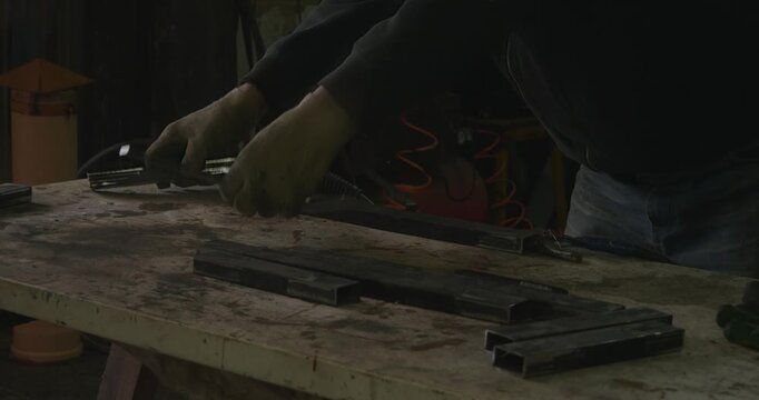 Unrecognizable craftsman wearing protective gloves carefully joining metal pipe sections together on a wooden workbench inside his dimly lit garage workshop before starting to weld them.