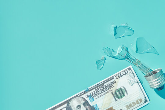 Broken lightbulb with scattered 100-dollar bills on turquoise background, wasted electricity and bankruptcy concept, copy space