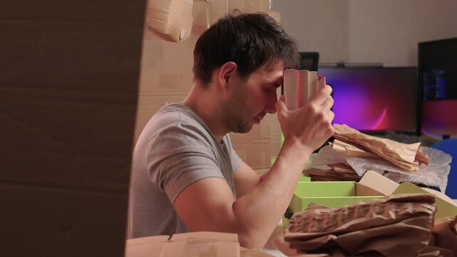 White man checking book for damage with worried expression, holding item to forehead under warm studio light amid torn kraft paper and monitor glow while deciding reship or return