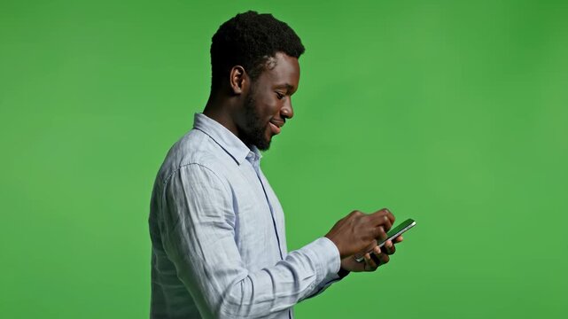 Happy African American man uses his mobile phone for social media or messaging, isolated on green screen. He is engaged and smiling while interacting with the device.