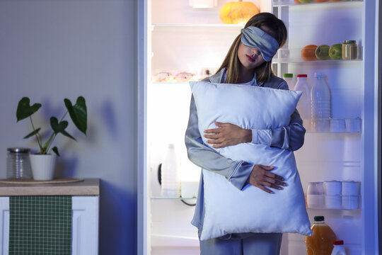 Female sleepwalker with sleeping mask and pillow near fridge in kitchen at night