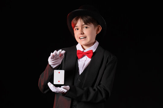 Young magician in tuxedo and top hat performing card trick on black background, smiling child illusionist in white gloves presenting playing card on stage