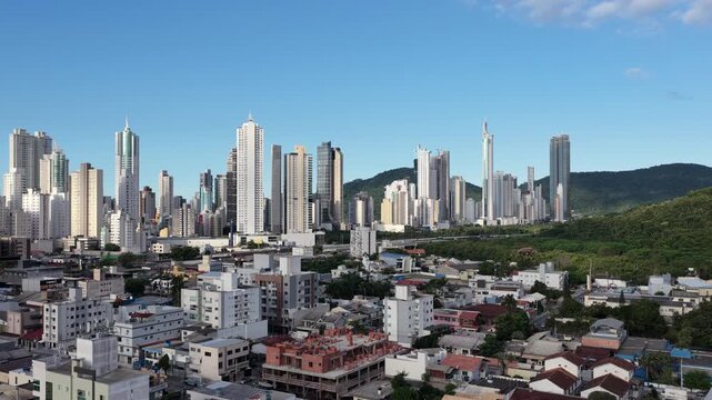 High-rise buildings dominate the skyline of Balne&aacute;rio Cambori&uacute;, surrounded by urban sprawl and lush hills. Ideal for cityscape features or urban development projects.
