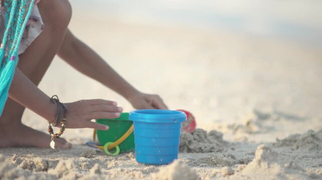 A girl with african braids in a summer costume plays on the beach with shells near the sea with waves under sunny sunset