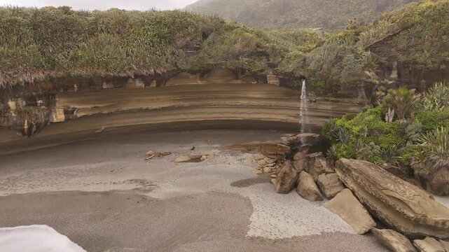 Unique waterfall cascades from stratified cliff directly onto Punakaiki sandy beach of Truman Track on New Zealand's West Coast. Aerial drone