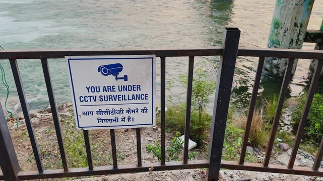 Close view of CCTV surveillance warning sign mounted on railing near riverbank, camera icon and text visible with flowing water and structure in background