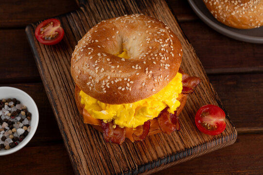 Bagel sandwich with crispy bacon, cheddar and scrambled eggs on a wooden board on a dark wooden background. Delicious breakfast. Rustic style.