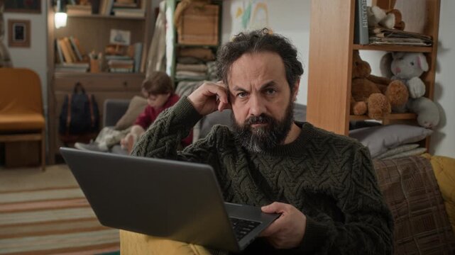 Portrait shot of gloomy middle aged bearded man sitting on couch with laptop while his son playing and wife cooking behind him in old run-down apartment