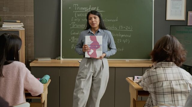 Medium full dolly-in shot of Hispanic female teacher talking to high school students about pregnancy, embryo development by trimesters while holding childbirth guide book at reproductive health lesson