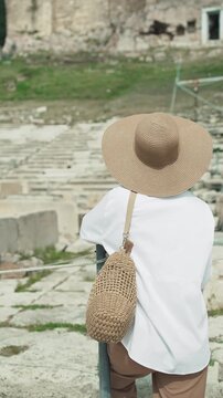 Tourist woman sith hat and glasses visiting Greek Theatre of Dionysus at the foot of Acropolis, Athens, Greece, cinematic isolated shot, vertical orientation