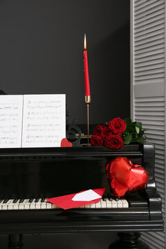 Romantic valentine still life with red roses, burning candle, love letter and heart balloon on piano keys with sheet music in elegant dark interior