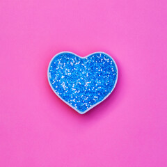 Fototapeta premium Bright blue glitter in a heart-shaped container sits on a pink background during daylight