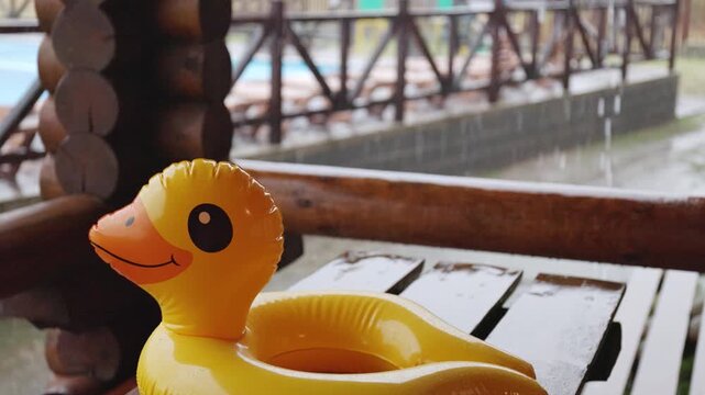 An inflatable duck lies on wooden table in gazebo at recreation center against the backdrop of pool covered with rain