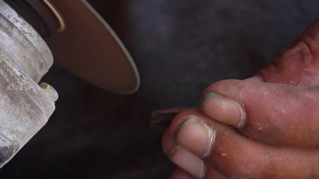 Close up handheld shot of a professional craftsman's dirty hands sharpening a metal drill bit using a power angle grinder. The footage captures the intense industrial process, showing the right hand h