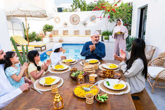 Arab family wearing traditional emirati clothing having lunch at home in the backyard garden - Middle-eastern family bonding and having fun on a family reunion dinner