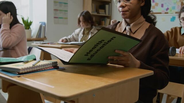 Medium shot of teenage African American girl opening textbook folder with lesson handouts and reading questions for test or assignment during health education lesson at middle school