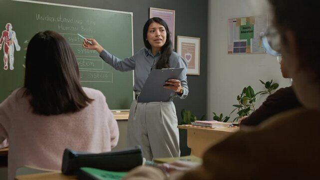 Over-shoulder rack focus shot of diligent male school student listening to Hispanic female teacher, who is standing by blackboard and explaining human body anatomy at health education lesson