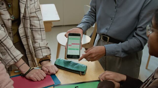 Medium close-up of Hispanic female teacher demonstrating menstrual tracker app on smartphone to group of teenage girls at middle school during candid discussion about reproductive health