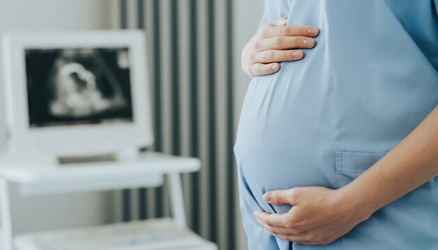 A pregnant woman in medical scrubs gently holds her belly during an ultrasound examination in a clinical setting.