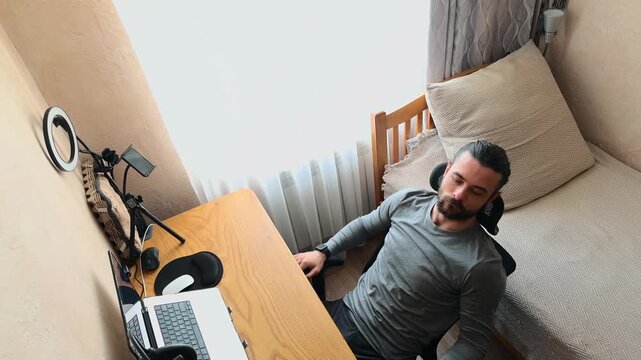 Recognizable Caucasian Young Man with Man Bun Sitting at Wooden Desk Leaning Back in Mesh Chair Thinking and Contemplating then Starting to Type on Laptop at Home Office Studio High Angle Perspective