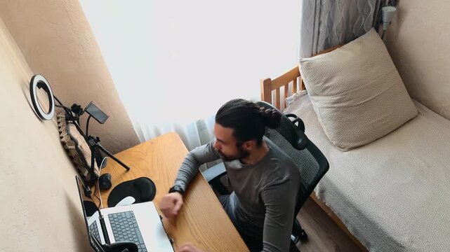 Young Caucasian Man with Bun Hair Entering Home Office Studio Sitting in Mesh Chair Stretching Arms and Hands then Starting to Type on Laptop at Wooden Desk High Angle Tilted View Authentic Day