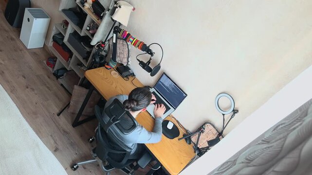 Young Caucasian Professional Man with Man Bun Typing on Laptop in Creative Home Office High Angle Tilted View Software Developer Programmer Coding at Wooden Desk with Professional Microphone Shot