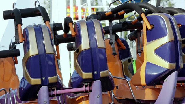 Close-up view of vibrant ride seats designed for an exhilarating amusement park attraction. The seats feature orange cushions and purple protective panels, equipped with black safety bars and straps. 