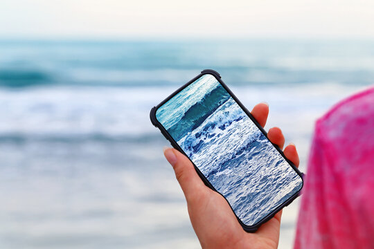 Person holds phone showing ocean waves while standing on sandy beach near water during daylight hours