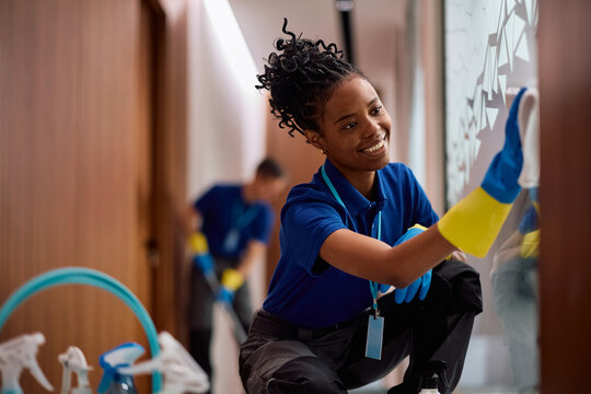 Happy black female cleaner wiping glass walls in office building.
