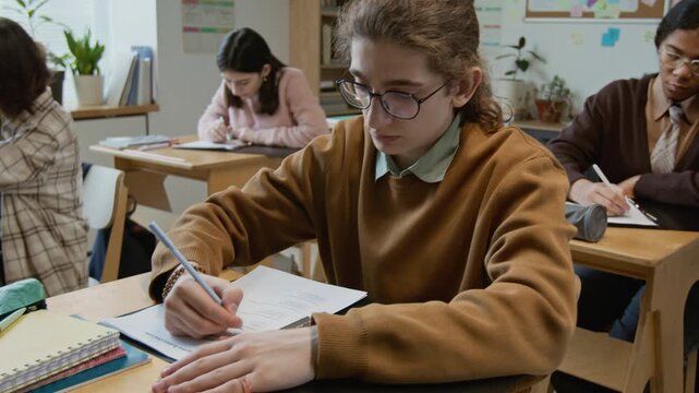 Medium full arc shot of diverse group of Caucasian and African American male and female high school students writing end of trimester test at lesson, focusing on task to achieve good academic grades