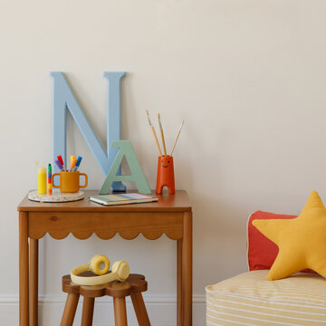 Stylish child room with wooden desk, colorful pens, brushes, and oversized letters under a mock up poster frame- bright and inspiring decor.