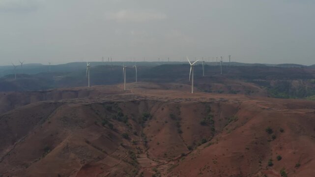 White wind turbines stand on eroded brown hillsides with visible gullies and sparse vegetation. Slow moving wide angle aerial shot captures the renewable energy installation across the ridgeline.