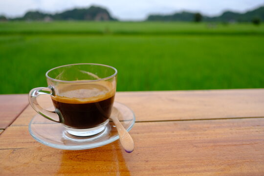 A cup of hot black coffee on a wooden table with beautiful green rice field and mountain background in the morning.
