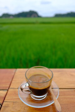 A cup of hot black coffee on a wooden table with beautiful green rice field and mountain background in the morning.