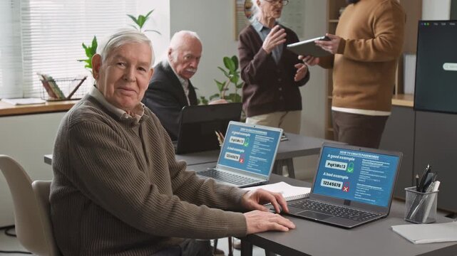 Medium shot of white haired senior man using laptop while sitting at desk in computer literacy class for seniors and turning looking at camera with smile, copy space