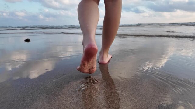 Close-up low angle view of a person walking bare feet on a wet sand beach with ocean waves and sky reflection.