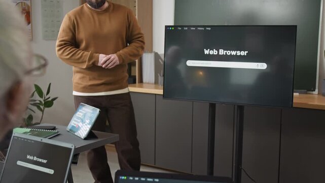 Tilt up shot of male teacher giving lecture in computer class for seniors and pointing at digital screen with web browser and search bar focus flowing from laptop on students desk, copy space
