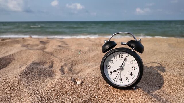 A classic black alarm clock sitting on a sandy beach with ocean waves in the background. Concept of vacation time, summer holiday, relaxation, and work life balance. 4K horizontal slow motion footage