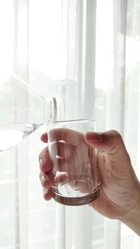 Hand pouring fresh pure water from a plastic bottle into a glass against a bright window background with natural light. Concept of healthy lifestyle, hydration, and clean drinking water.