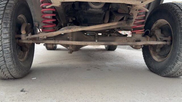 Close-up crossover suspension undercarriage view dusty asphalt gravel ground, red coil springs and muddy axle visible, worn tires