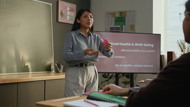 Medium full shot of young Hispanic female teacher demonstrating menstrual cup to class of schoolgirls, informing about its function and convenience during reproductive health lesson