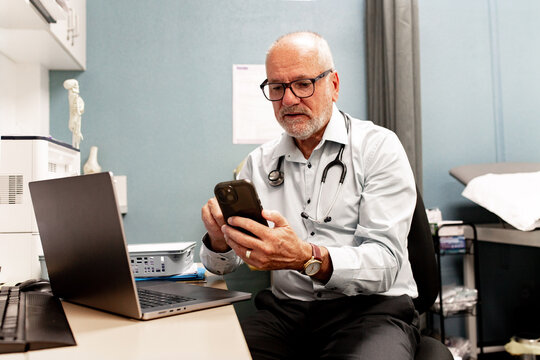 Male medical professional using smartphone with an open laptop in clinic