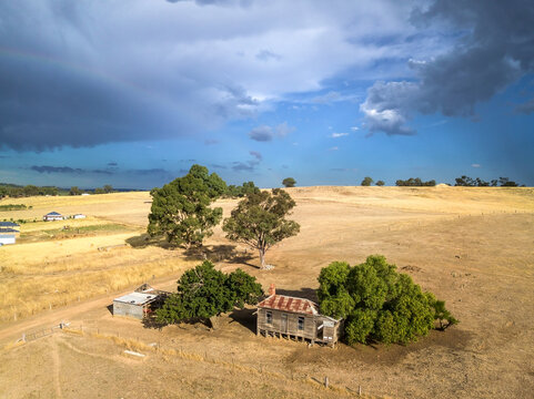 Aerial view of an abandoned farmhouse in a dry paddock under a dramatic sky and faint rainbow