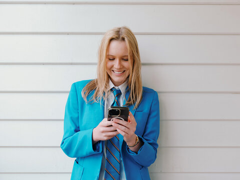 Teenage girl in school uniform leaning against the white wall while engaged on her phone