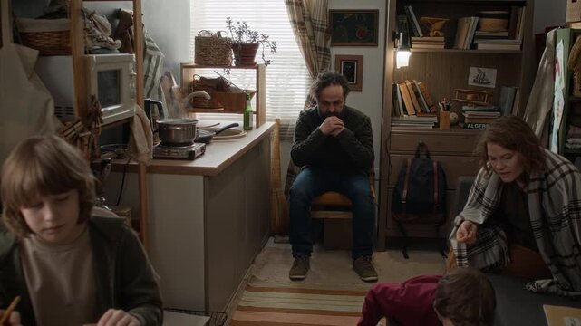 Handheld zoom in shot of depressed man drinking beer while his son doing homework and his mother and young brother playing on couch in small, rundown apartment.